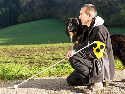 Ein Mann kniet mit einem Blindenstock neben einem Hund in einer Landschaft.