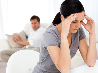 Eine gestressete Frau hält sich den Kopf, während ein Mann im Hintergrund auf einem Sofa sitzt.